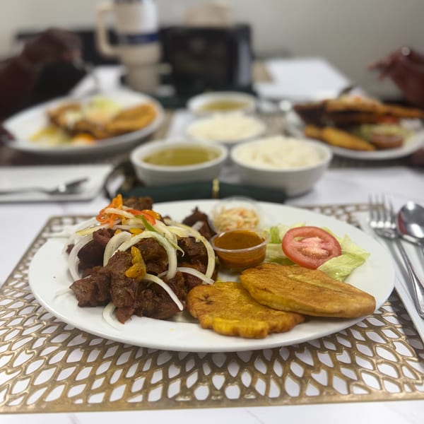 A plate of fried meat with onions, plantains, salad, and sauce, with other dishes blurred in the background.