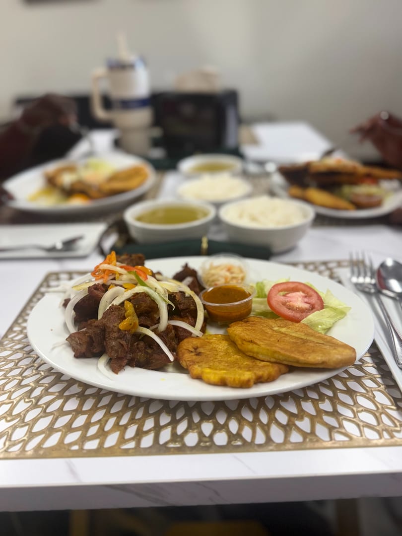A plate of fried meat with onions, plantains, salad, and sauce, with other dishes blurred in the background.
