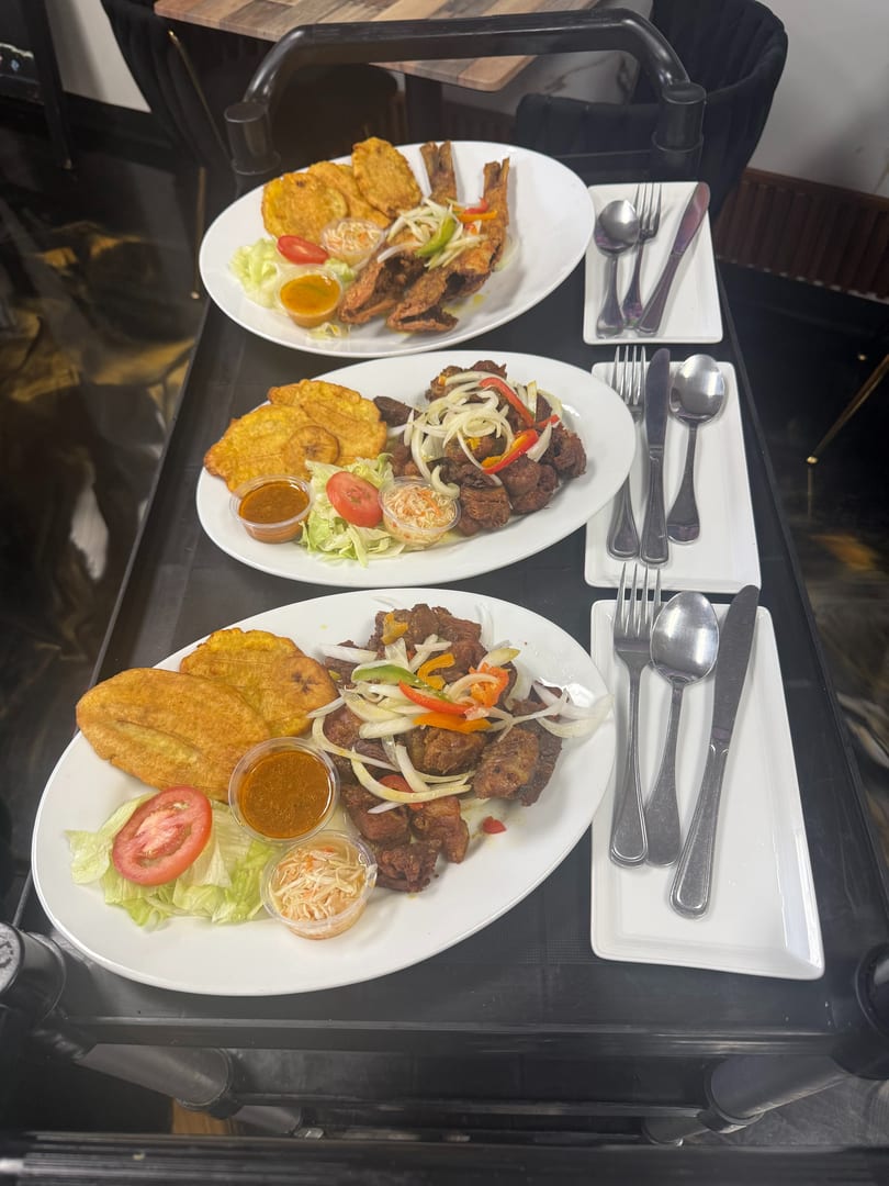Three plates of Haitian food with fried plantains, meat, salad, and sauces on a serving cart.
