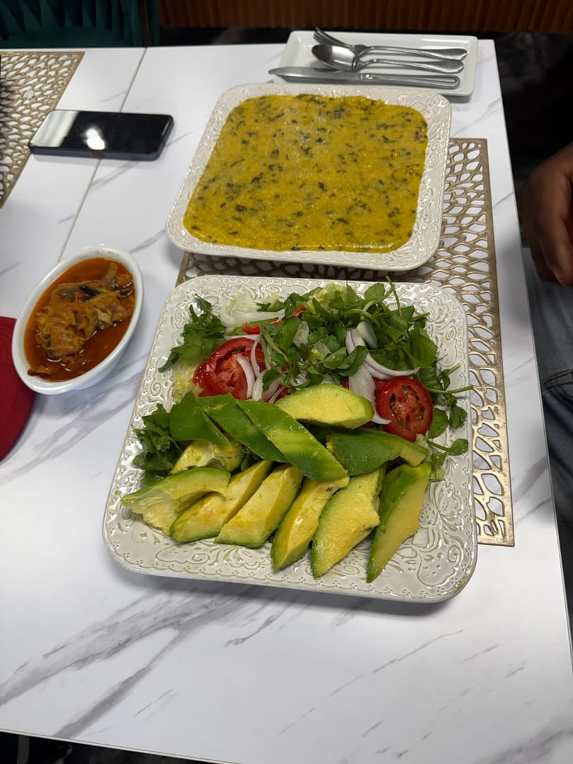 A table set with a vibrant green salad, sliced avocado, a yellow dish, and a bowl of stew.