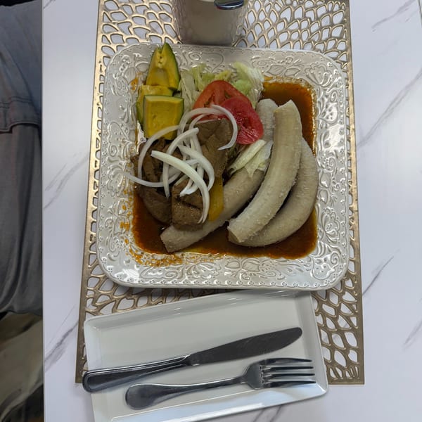 A plate of meat, plantains, avocado, and salad with a drink on a white marble table.