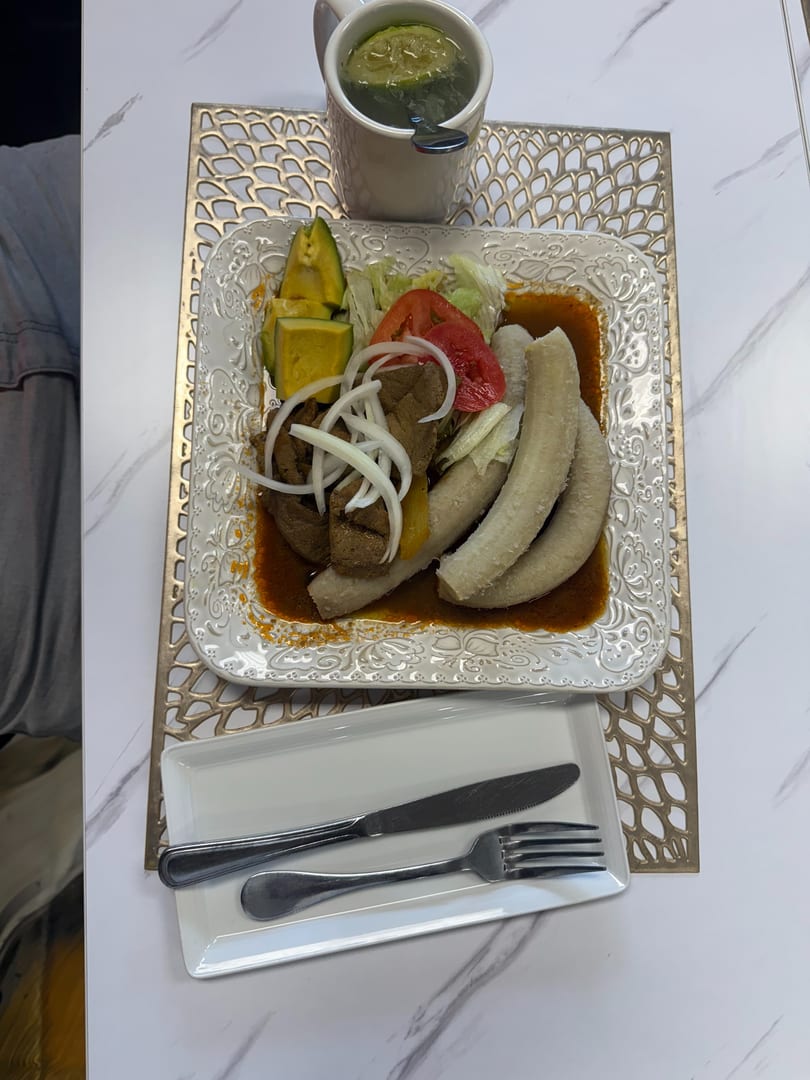 A plate of meat, plantains, avocado, and salad with a drink on a white marble table.